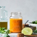 Side view of a jar of bright orange roasted red pepper dressing next to a cut lime and a bowl of salad greens