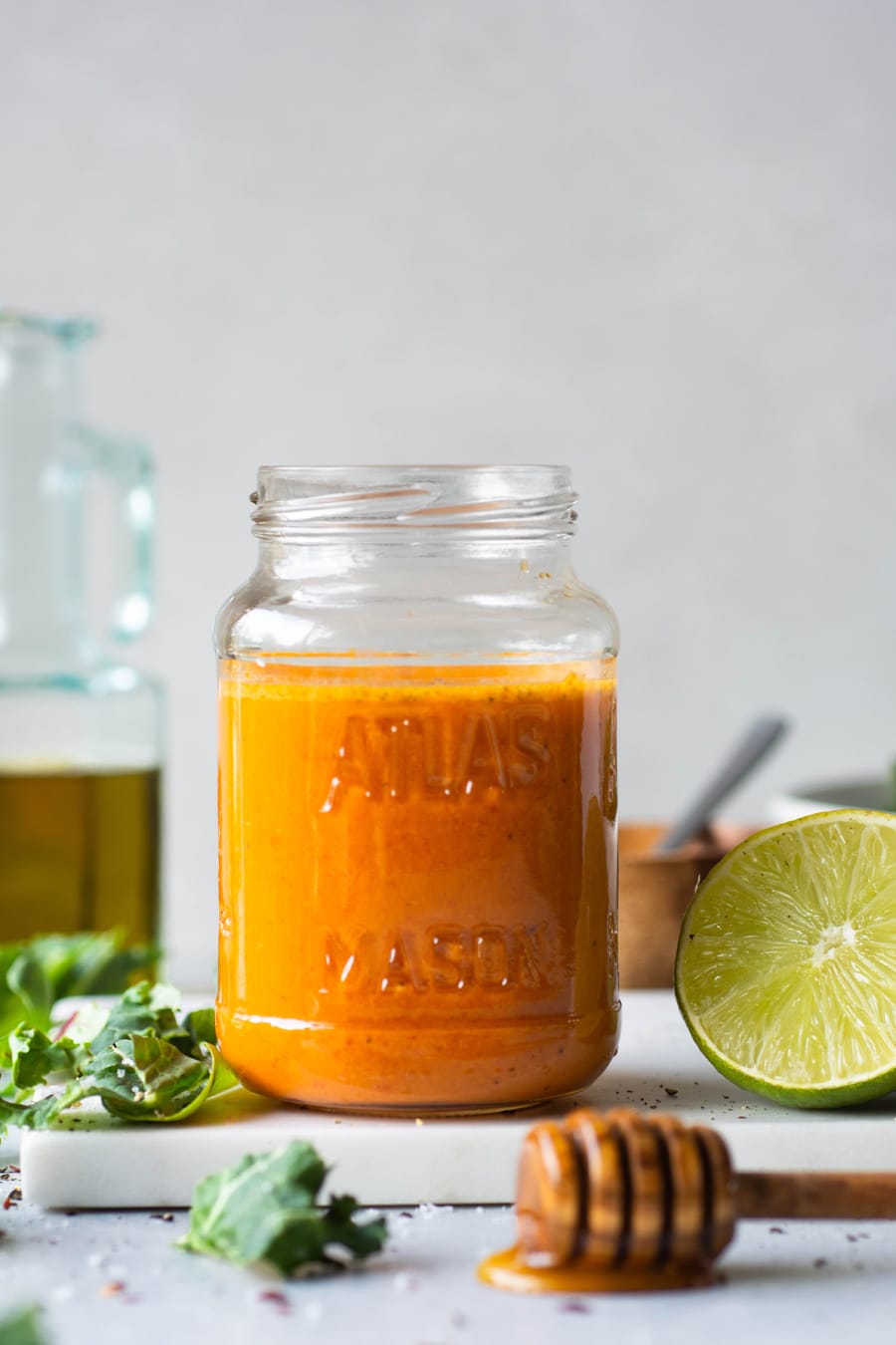 Side view of a jar of bright orange roasted red pepper dressing next to a cut lime and a bowl of salad greens