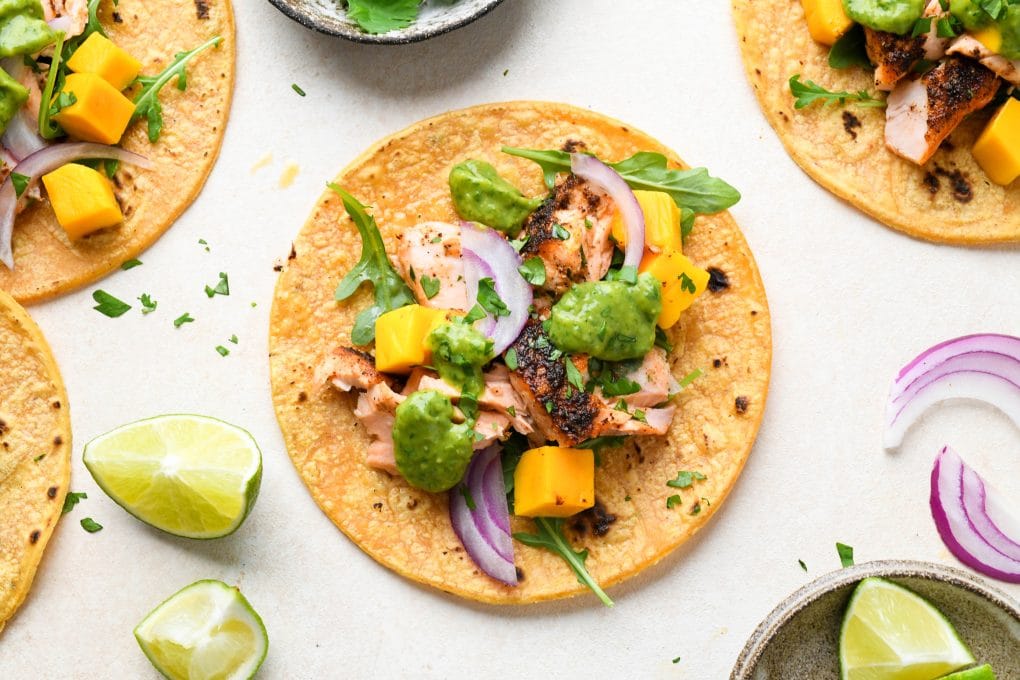 Several blackened salmon tacos on a light cream background surrounded by lime wedges and fresh cilantro.