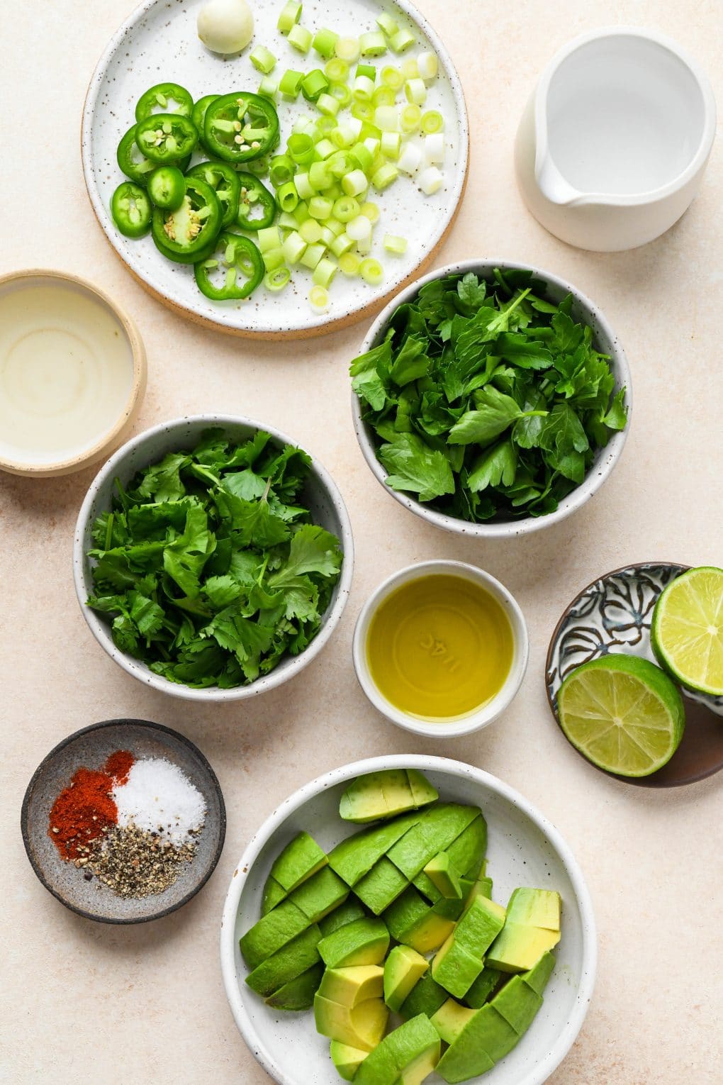 Ingredients for Avocado Herb Green Sauce in various ceramics, on a light cream colored background.