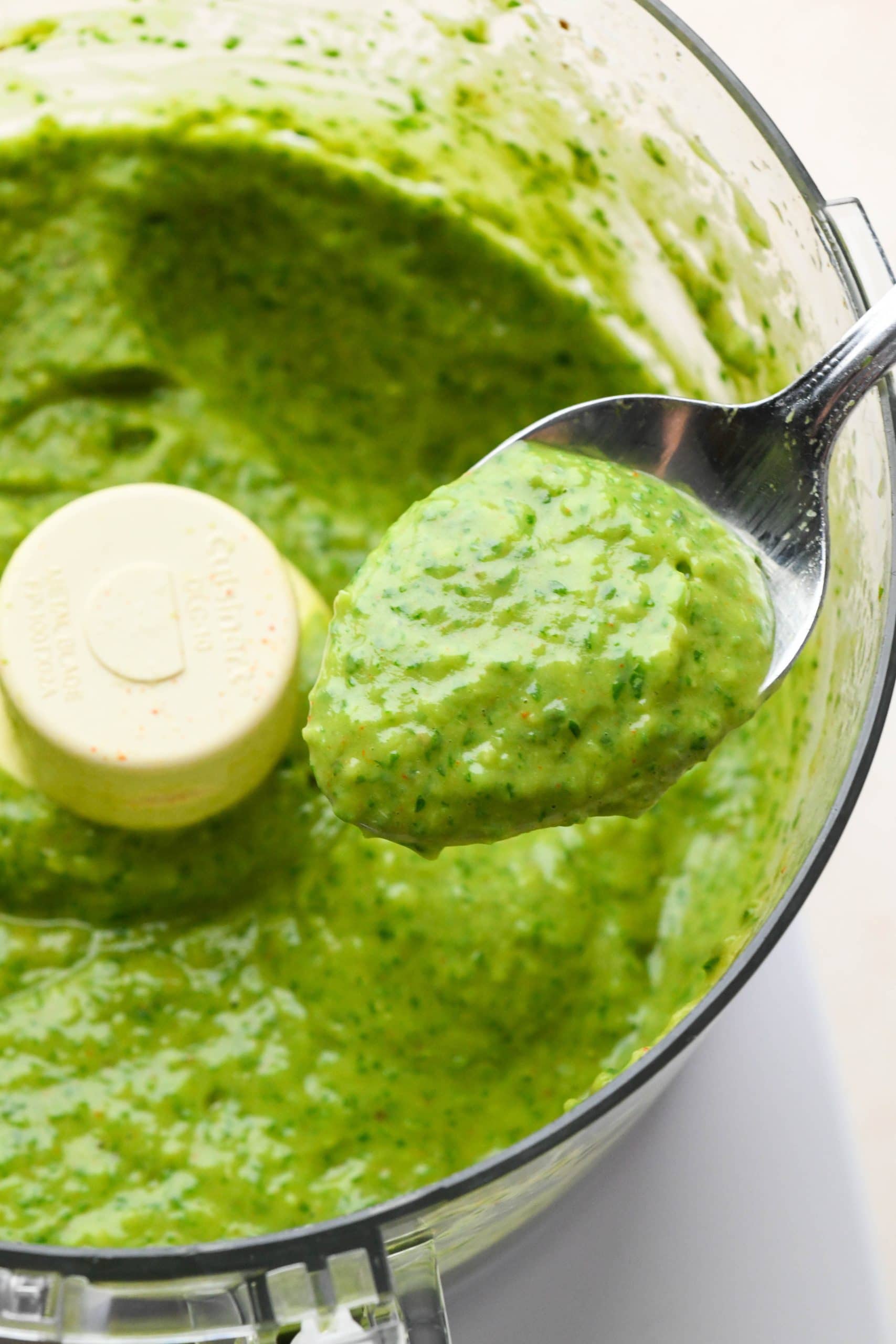 How to make avocado herb green sauce: A spoon lifting out a small amount of sauce from the food processor to show the creamy texture.