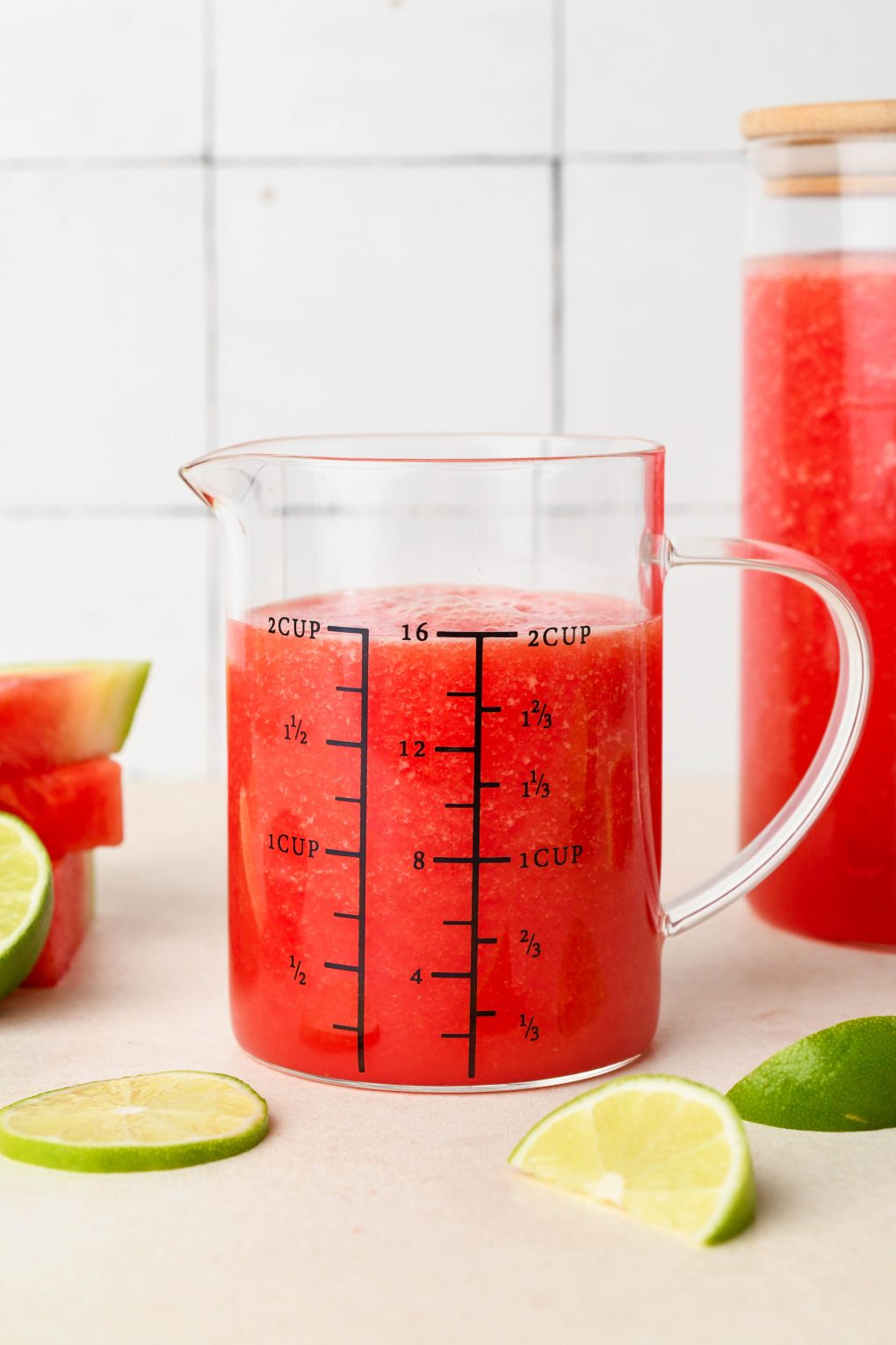Watermelon juice in a cute measuring glass, next to a larger jar also filled with juice. On a white background surrounded by lime wedges.