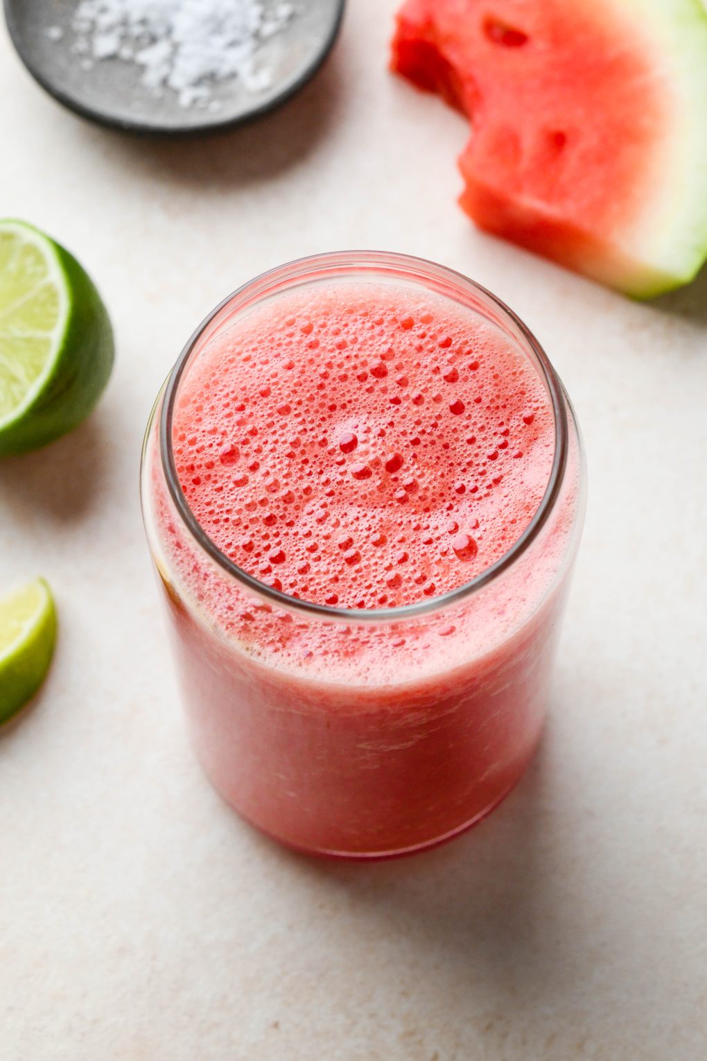 Watermelon juice in a cute glass on a light cream colored background, surrounded by cut watermelon wedges and cut limes.