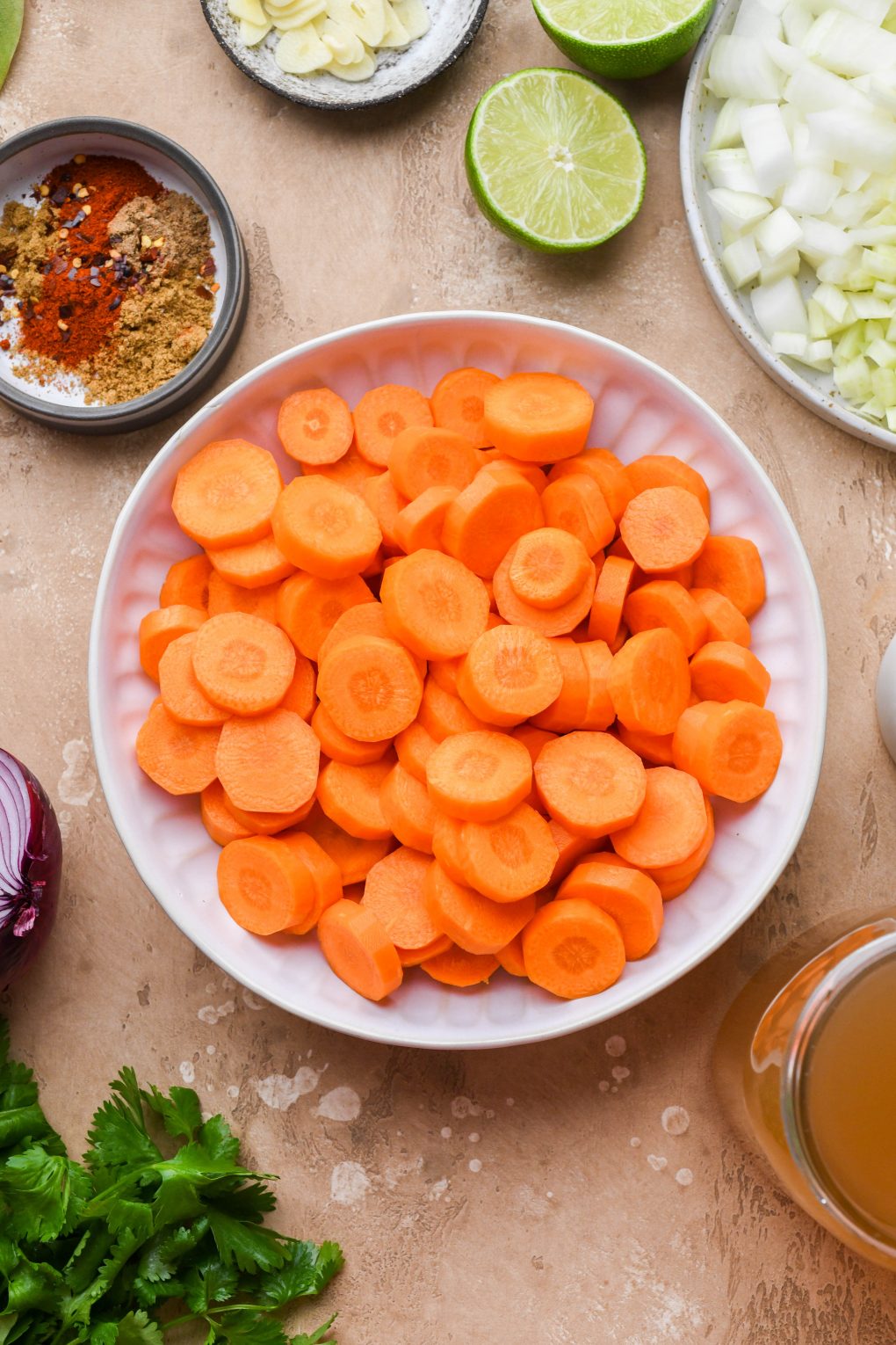 Image of ingredients for creamy carrot soup - sliced carrots in a large bowl, diced onion and fennel, chopped garlic, spices, red onion, cilantro, broth, and limes. On a light brown background.