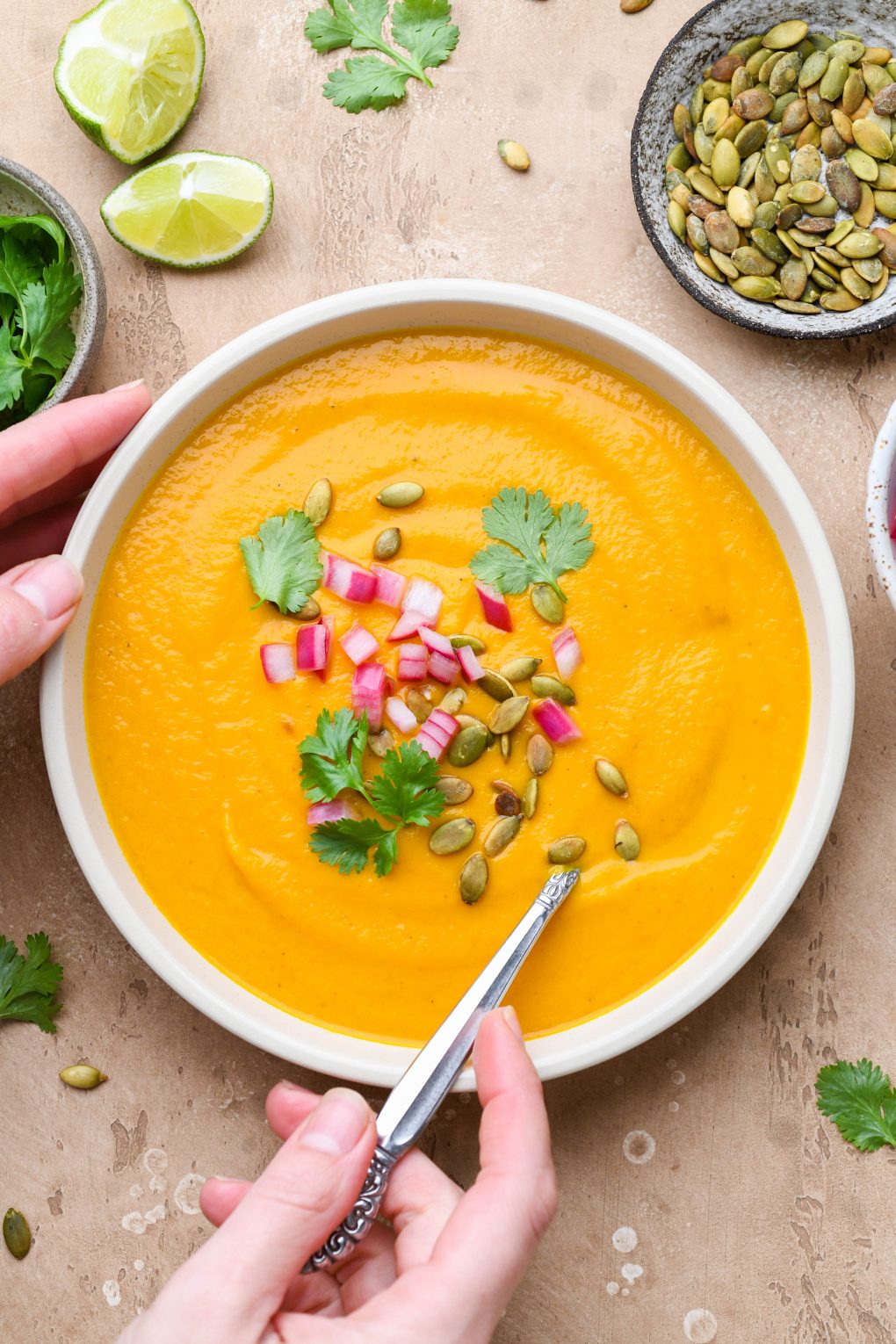 Large wide bowl of bright orange creamy carrot soup on a light brown background. Soup is topped with fresh cilantro leaves, chopped pickled red onions, and pumpkin seeds. Hands are reaching in to grab the spoon and bowl.