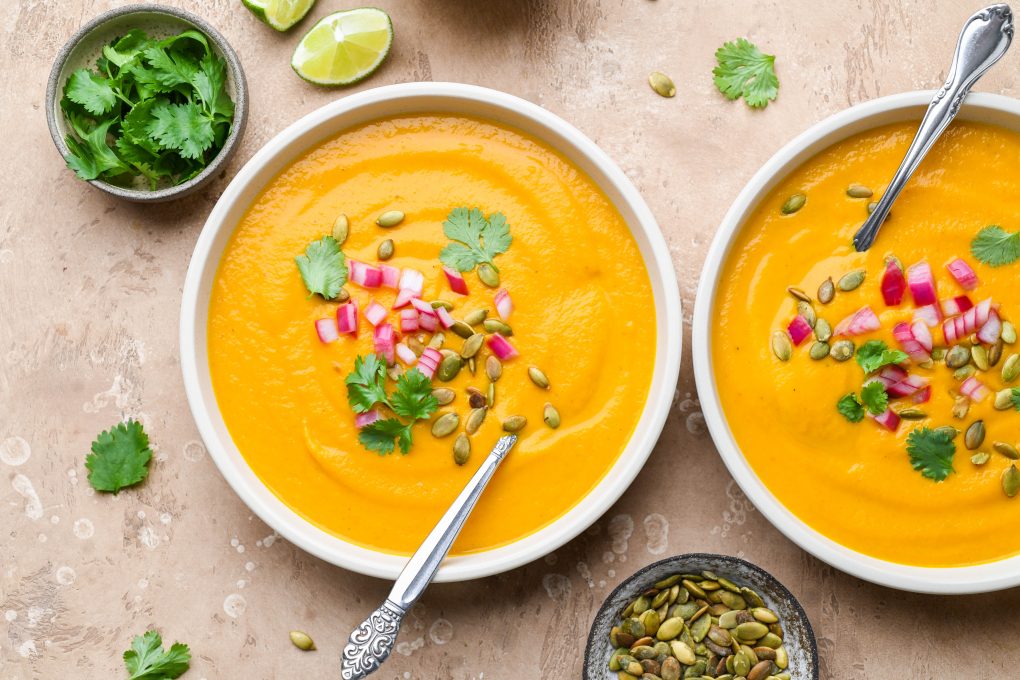 Two side by side wide bowls of bright orange creamy carrot soup on a light brown background. Soup is topped with fresh cilantro leaves, chopped pickled red onions, and pumpkin seeds.