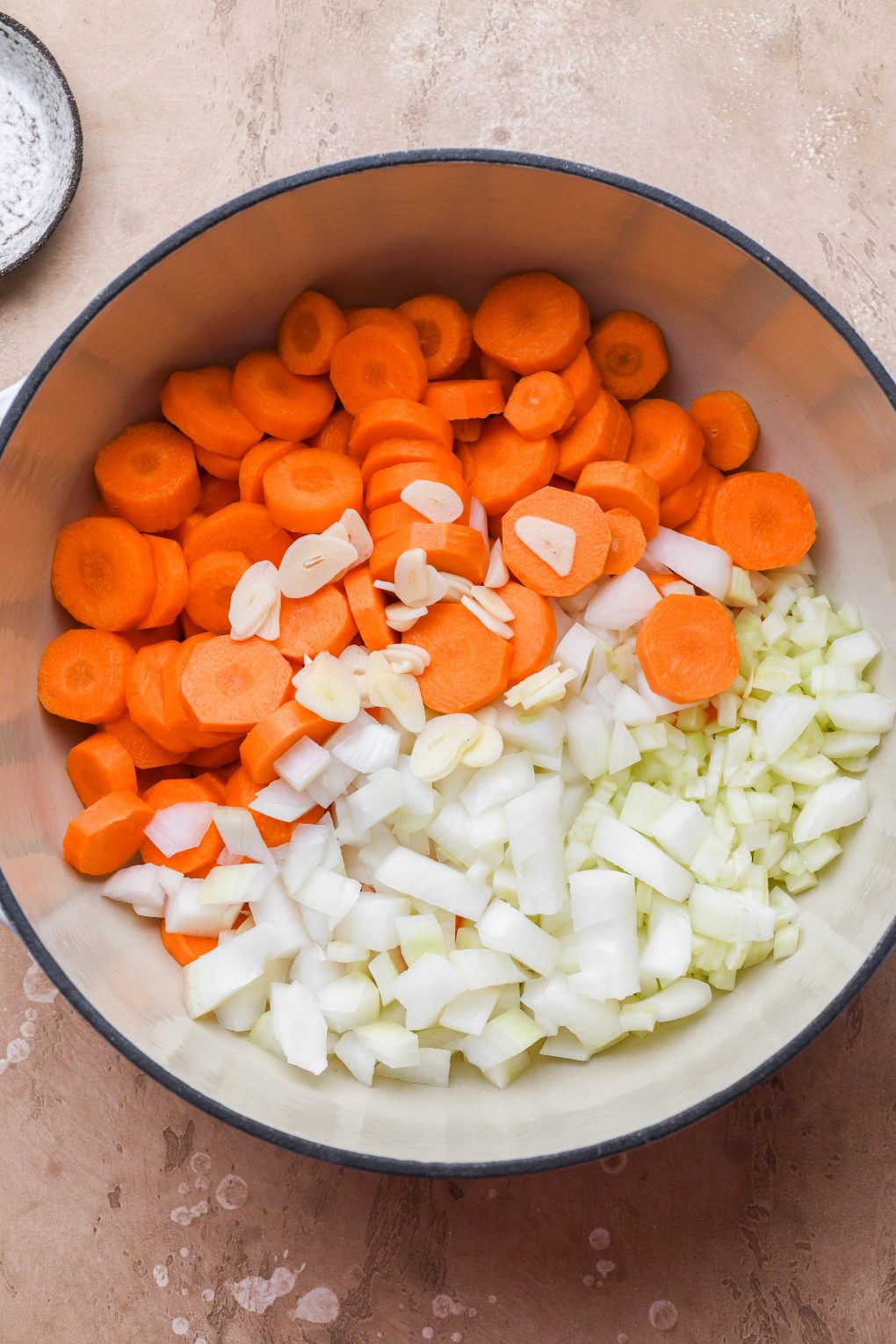 Chopped carrots, onions, fennel, and garlic in a large white ceramic soup pot. On a light brown background.