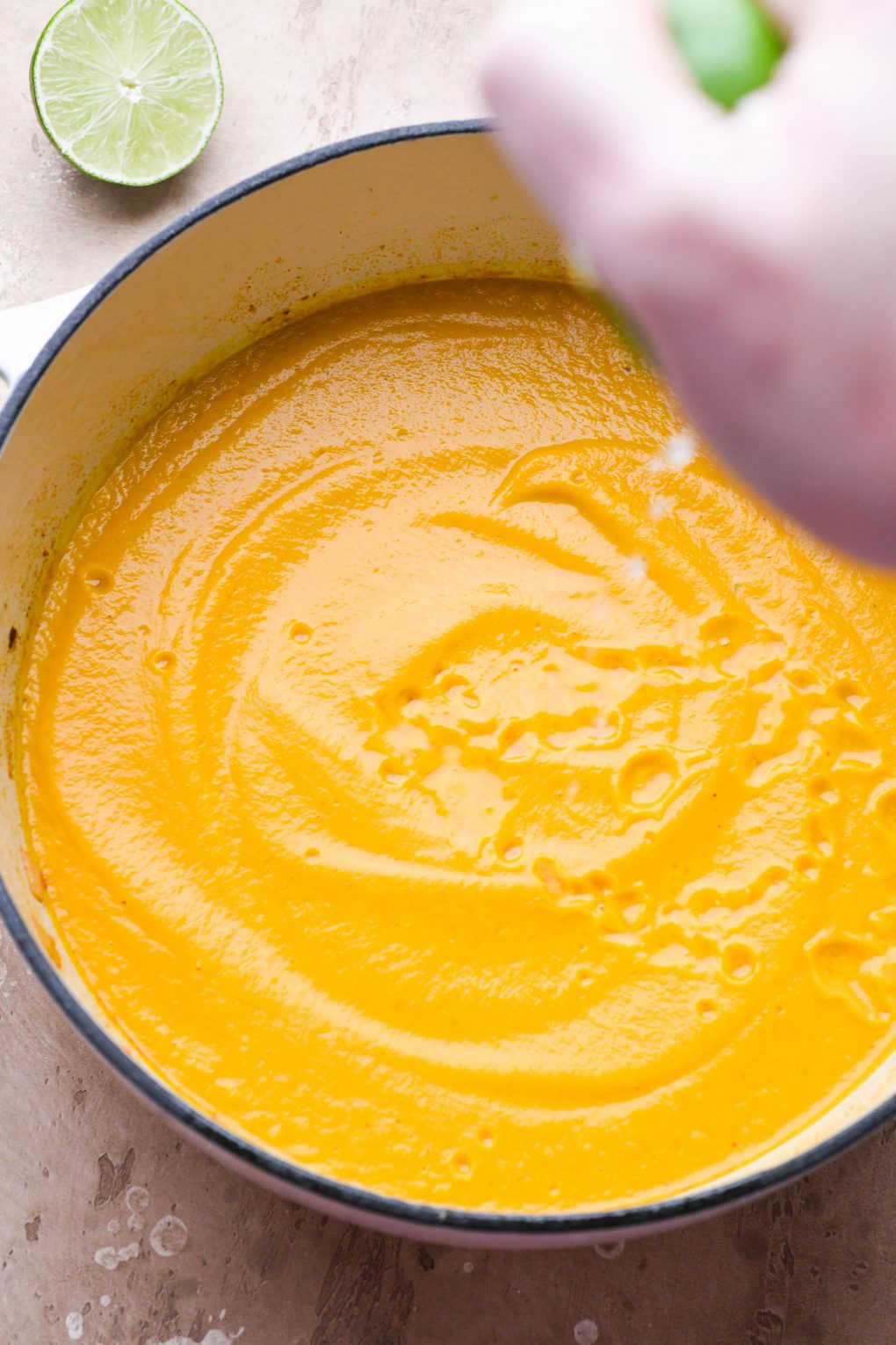 Hand squeezing lime into a large ceramic soup pot filled with creamy blended carrot soup.