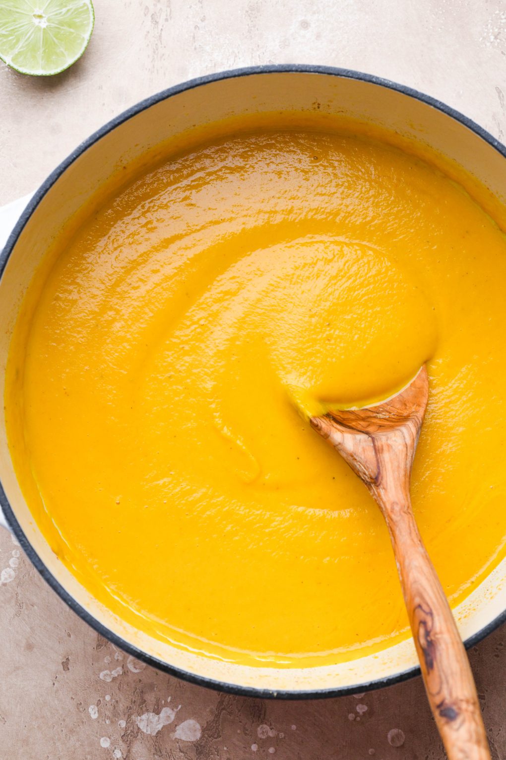Large wooden spoon dipped into a large ceramic soup pot filled with creamy blended carrot soup.