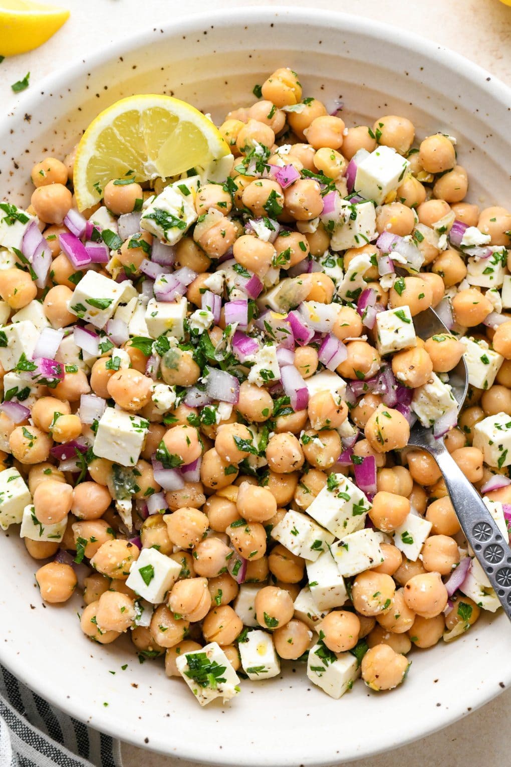 A large ceramic bowl of lemony chickpea feta salad with a lemon wedge and a spoon dipped into the salad.