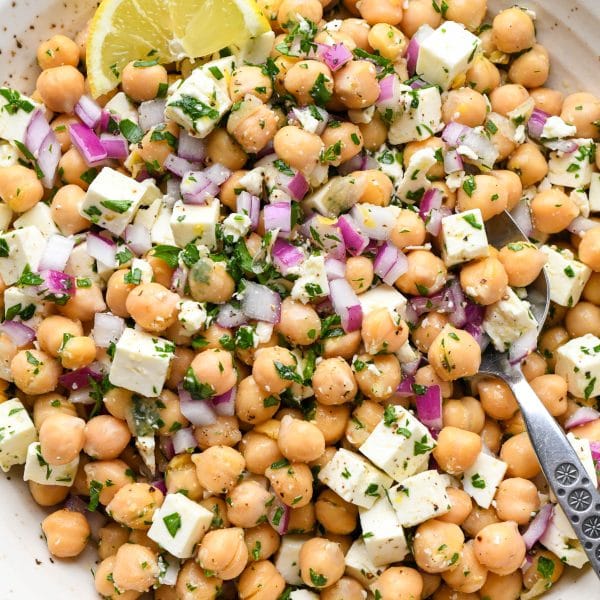 A large ceramic bowl of lemony chickpea feta salad with a lemon wedge and a spoon dipped into the salad.