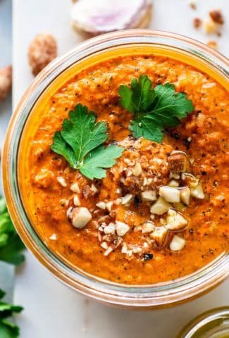 An overhead view of bright orange romesco sauce topped with parsley leaves, chopped almonds, and black pepper in a glass jar on top of a marble board