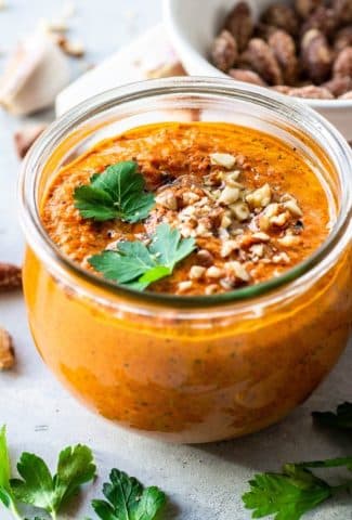 45 degree angle view of a clear glass jar of roasted red pepper romesco sauce topped with parsley, chopped almonds, and olive oil. On a white background with a bowl of nuts in the background.