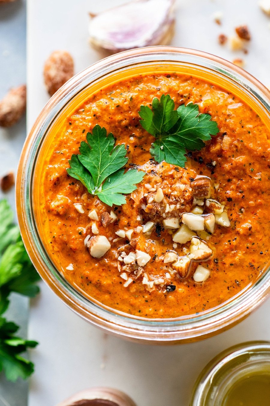 An overhead view of bright orange romesco sauce topped with parsley leaves, chopped almonds, and black pepper in a glass jar on top of a marble board