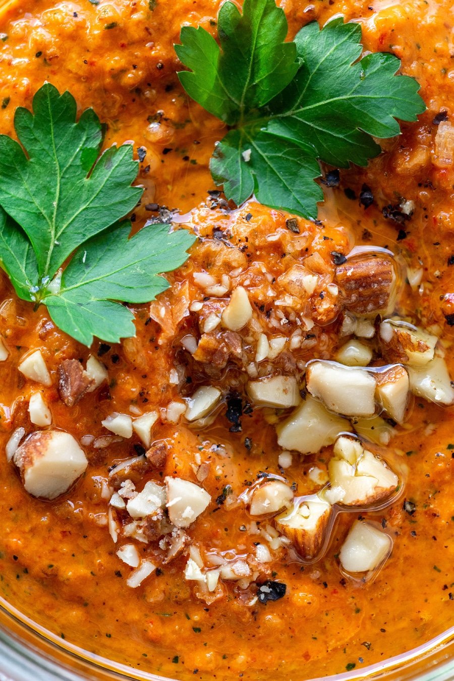A close up view of bright orange romesco sauce topped with parsley leaves, chopped almonds, and black pepper