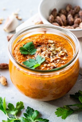 45 degree angle view of a clear glass jar of roasted red pepper romesco sauce topped with parsley, chopped almonds, and olive oil. On a white background with a bowl of nuts in the background.
