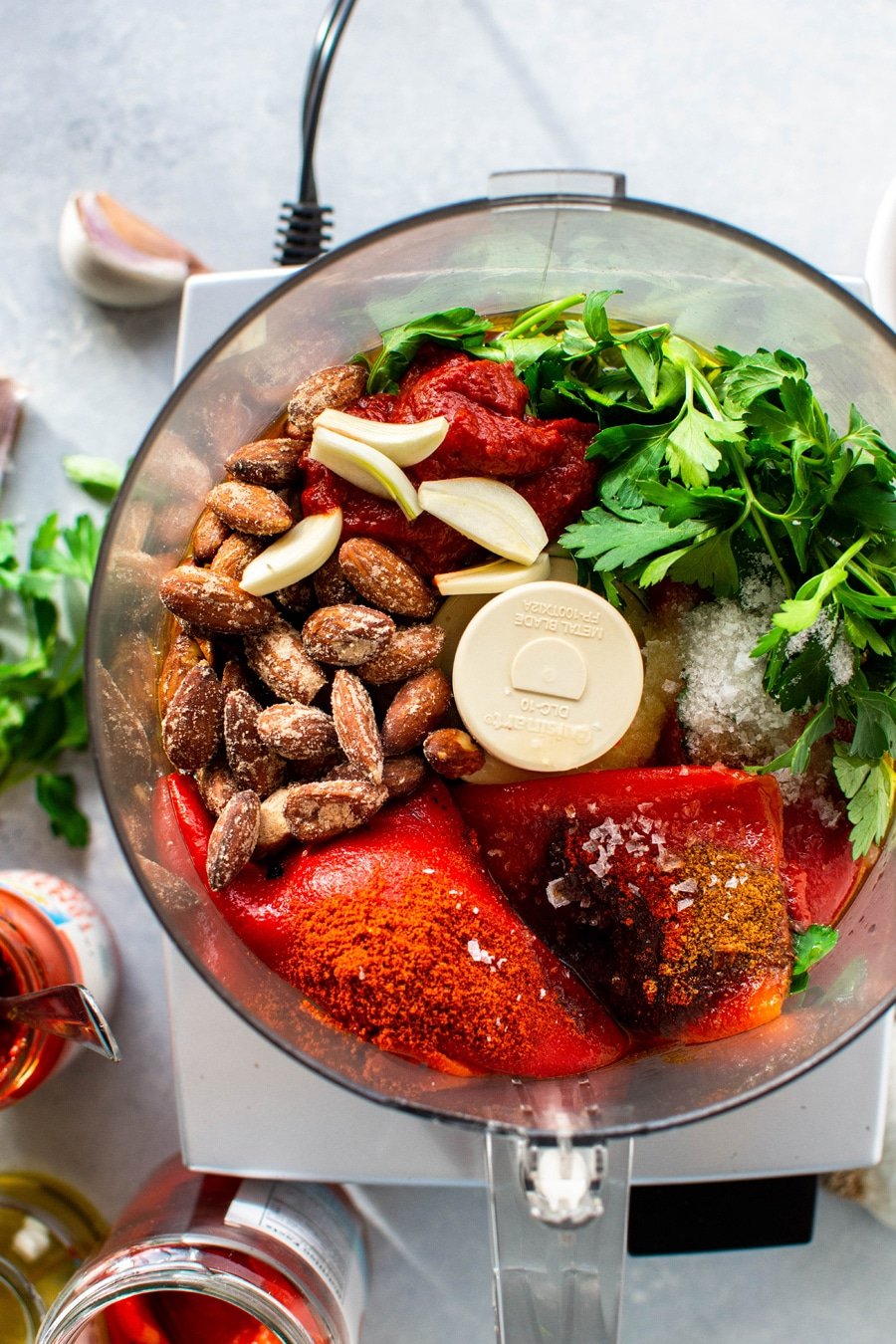 Overhead view of a food processor filled with all the ingredients for this fast and easy romesco sauce - roasted red peppers, almonds, garlic, parsley, spices, and salt.