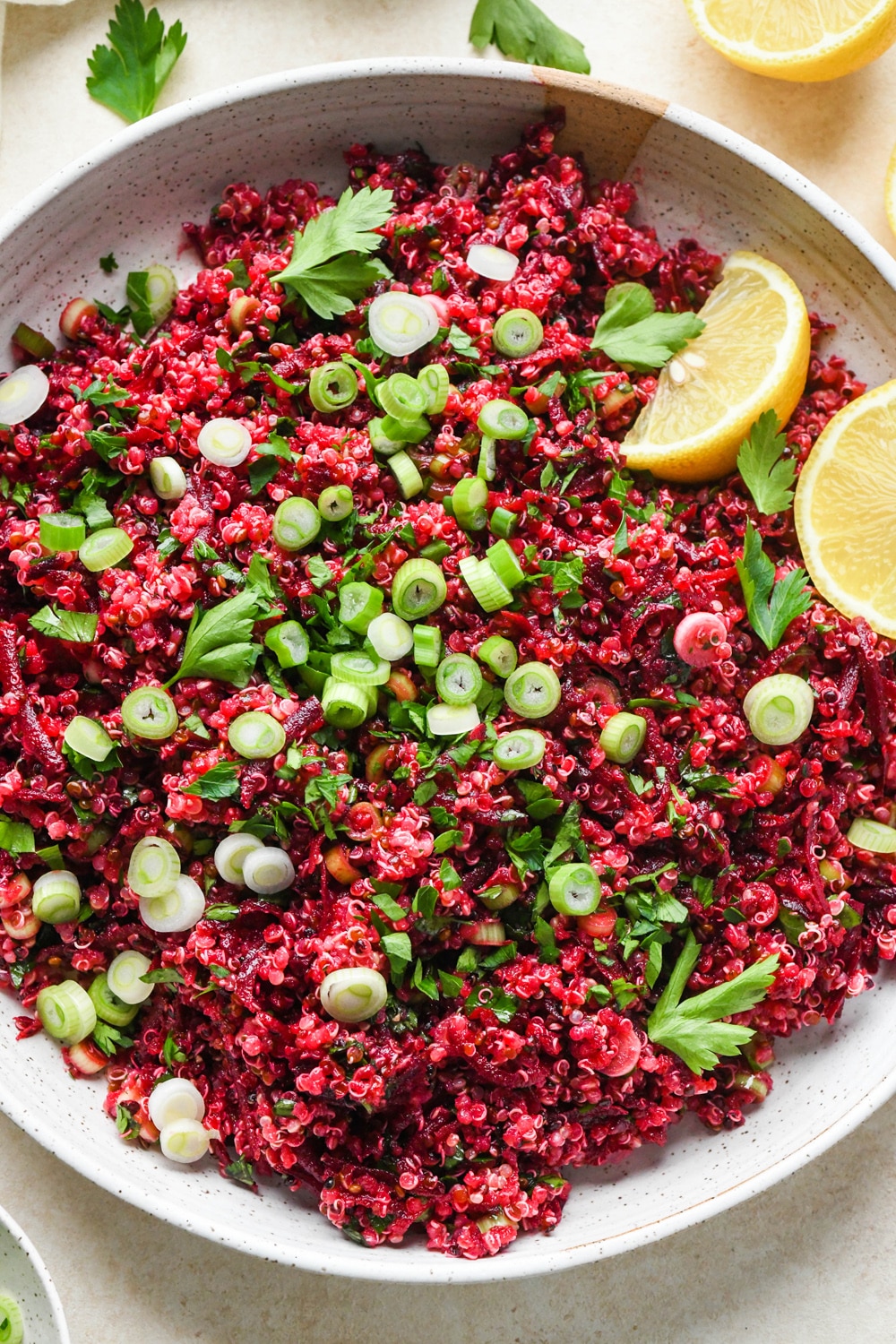 How to make quinoa and fresh beet salad: Deep pink quinoa salad in a large ceramic serving dish after garnishing with thinly sliced green onions, fresh parsley, and lemon wedges.