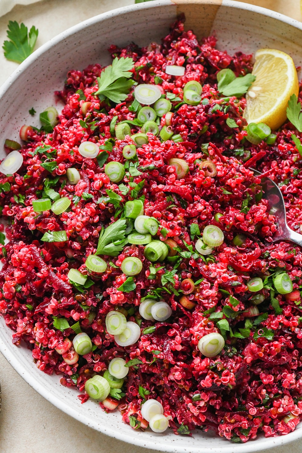 Quinoa and beet salad in a large shallow ceramic serving dish. The salad is garnished with sliced green onion, fresh chopped parsley, and lemon wedges on the side.