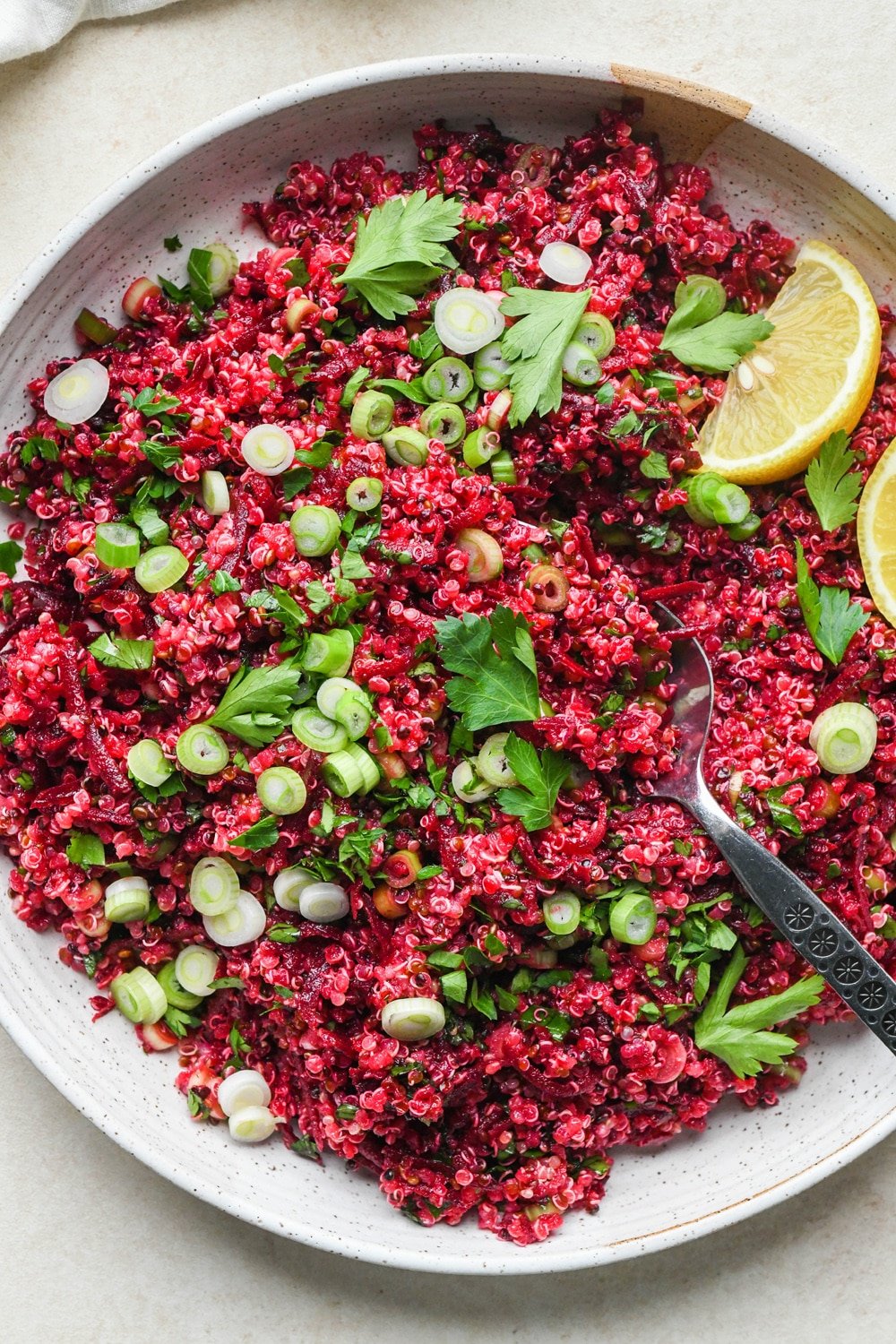 Quinoa and beet salad in a large shallow ceramic serving dish. The salad is garnished with sliced green onion, fresh chopped parsley, and lemon wedges on the side.