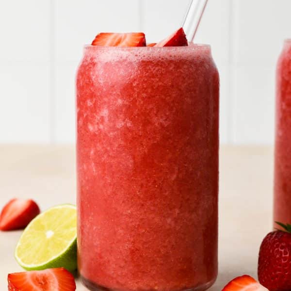Two glasses filled with strawberry hibiscus slushy, next to some fresh strawberries and cut lime, on a light beige background.