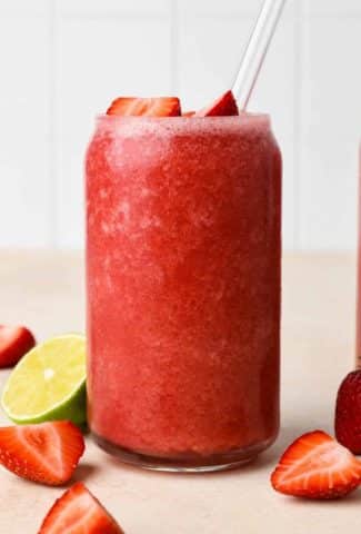 Two glasses filled with strawberry hibiscus slushy, next to some fresh strawberries and cut lime, on a light beige background.