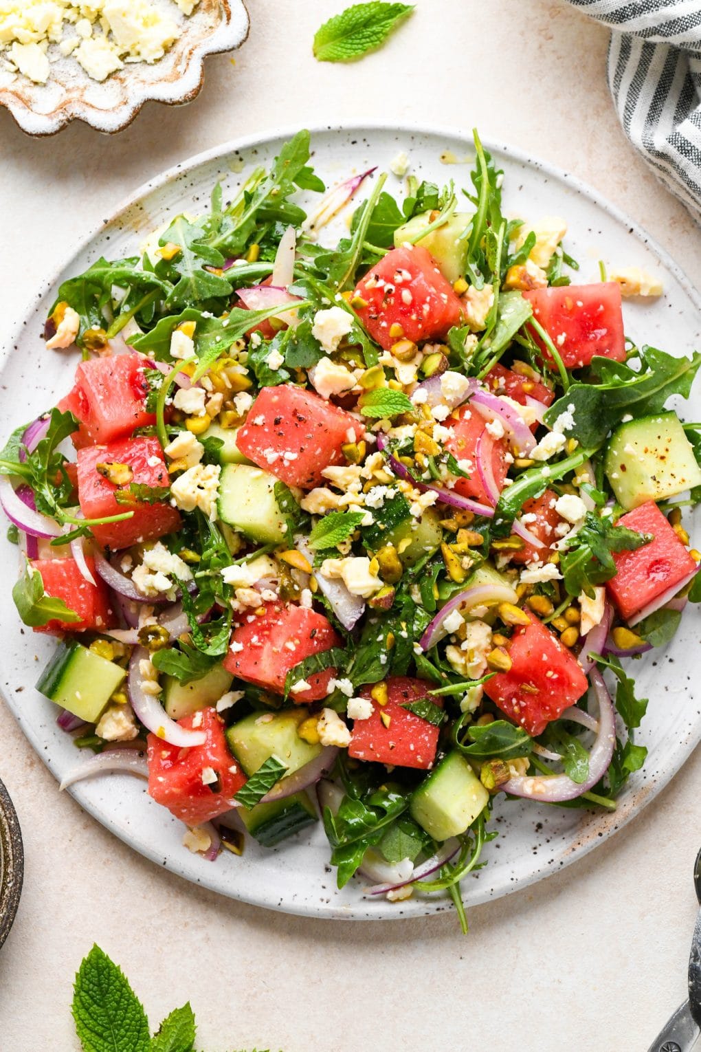 Watermelon and arugula salad with cucumber, feta, mint, and pistachios, piled high on a large white platter.