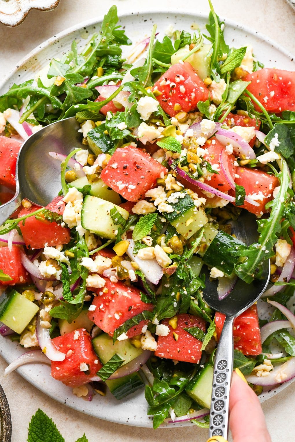 Watermelon and arugula salad with cucumber, feta, mint, and pistachios, piled high on a large white platter, with two spoons lifting some out to serve.