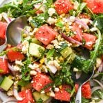 Watermelon and arugula salad with cucumber, feta, mint, and pistachios, piled high on a large white platter, with two spoons lifting some out to serve.