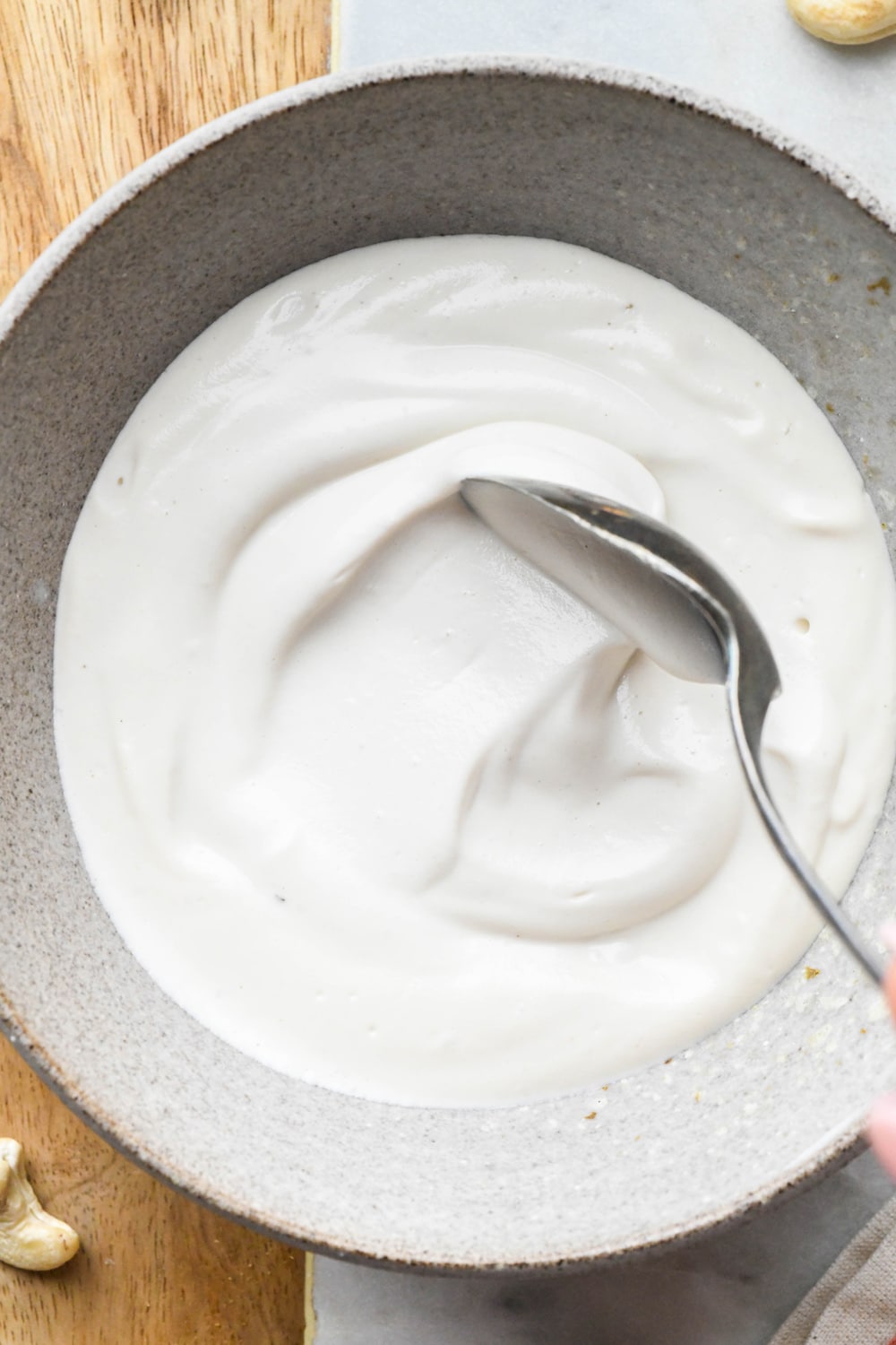 Cashew cream made with 1/2 cup of water in a grey ceramic bowl with a spoon stirring the cream to show the texture.