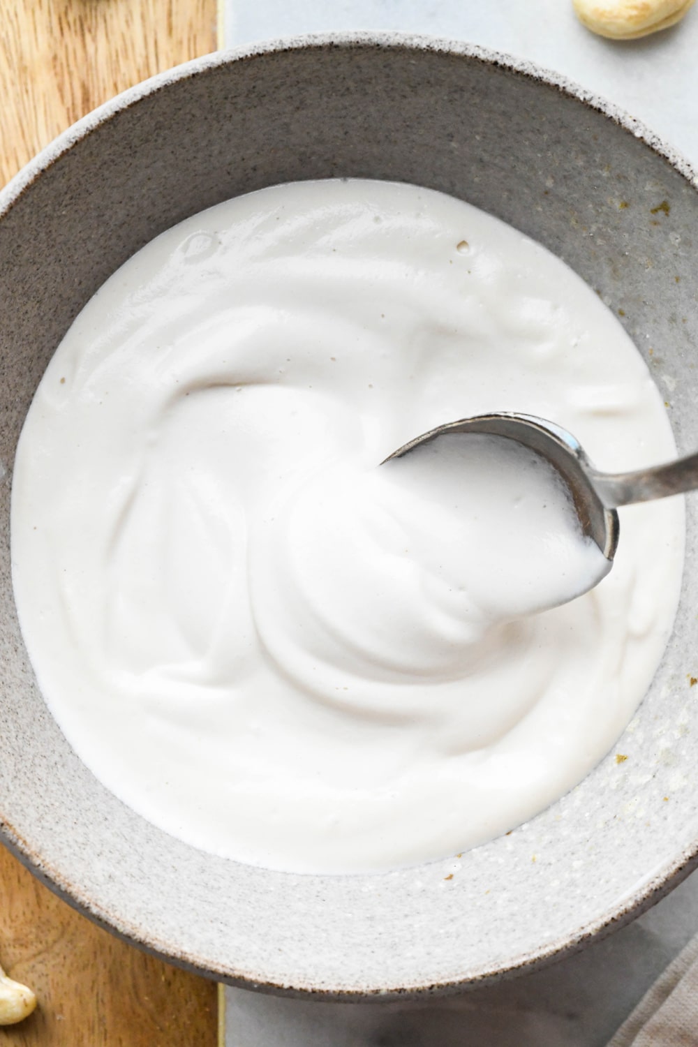 Cashew cream made with 1/2 cup of water in a grey ceramic bowl with a spoon lifting out some of the cream to show the texture.