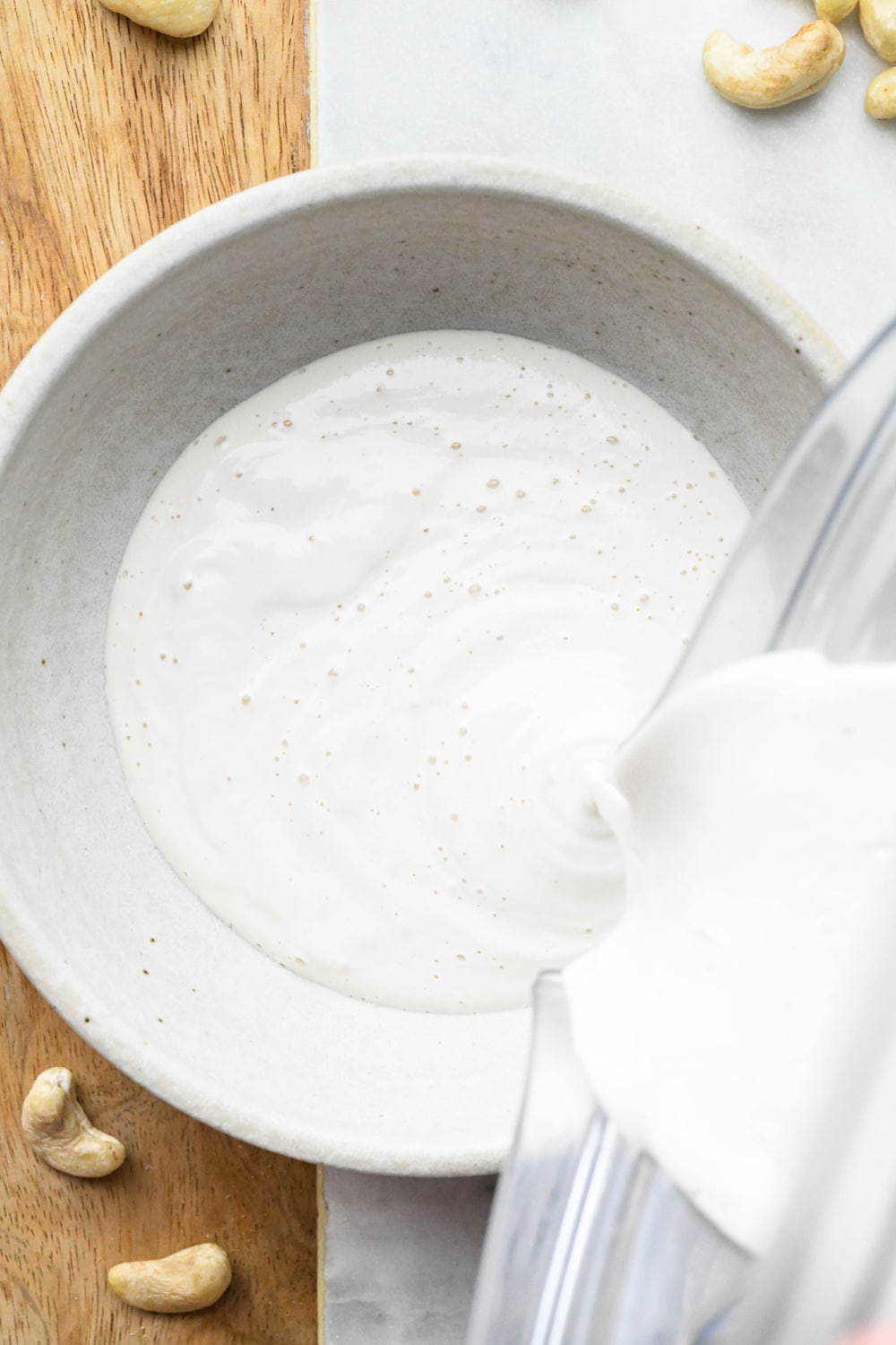A blender pouring cashew cream made with 3/4 cup of water into a grey ceramic bowl. 