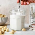 A hand lifting out a spoonful of cashew cream from a small jar surrounded by a dish and some scattered raw cashews.