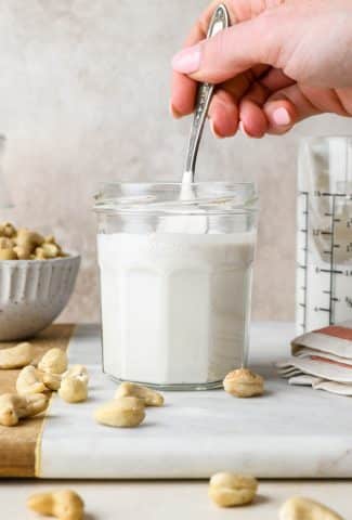 A hand lifting out a spoonful of cashew cream from a small jar surrounded by a dish and some scattered raw cashews.