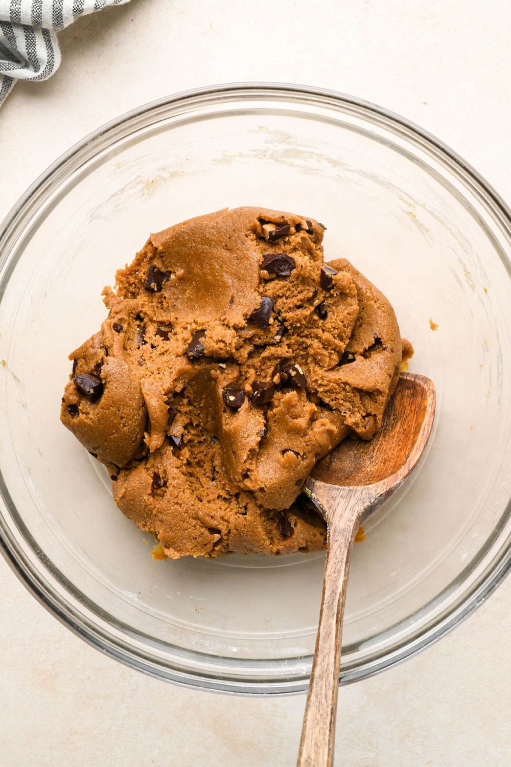 How to make Dark Chocolate Espresso Chickpea Flour Cookies: Chopped chocolate incorporated into cookie dough in a large glass bowl with a wooden spoon angled into the dough. 