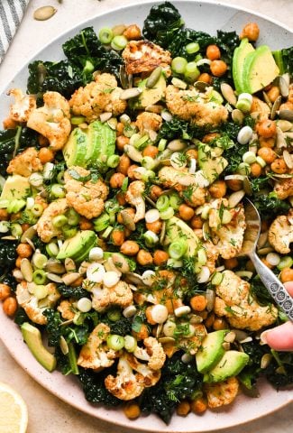 Chopped kale salad with roasted cauliflower and chickpeas on a large serving platter, garnished with thinly sliced green onions, and fresh dill. A serving spoon is lifting out a portion of the salad.