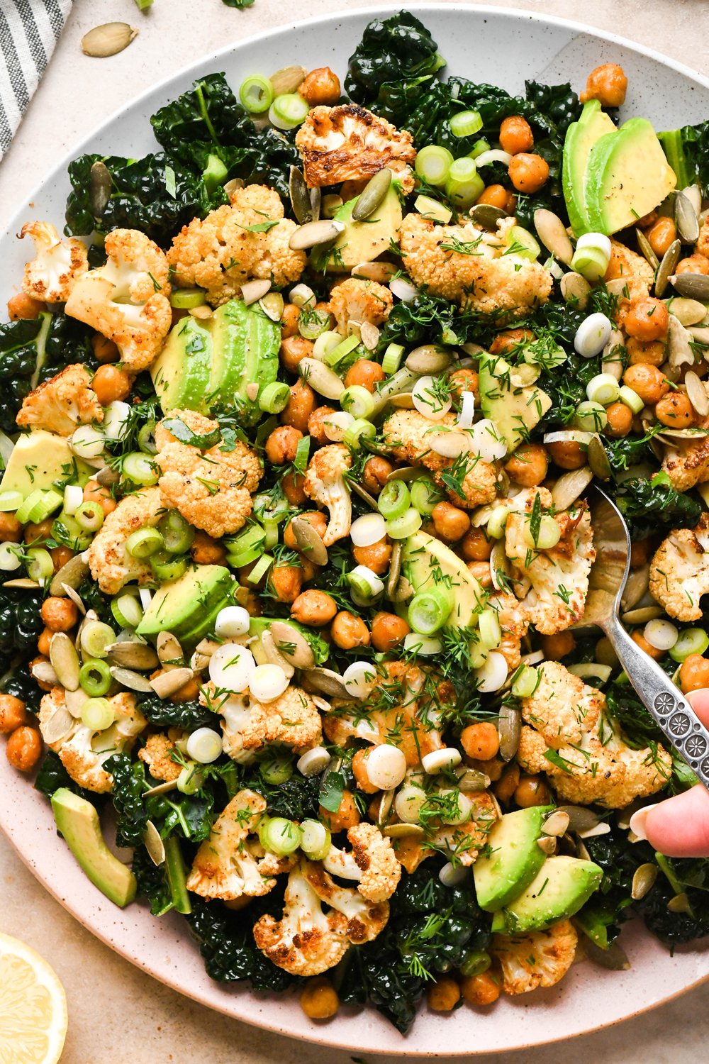 Chopped kale salad with roasted cauliflower and chickpeas on a large serving platter, garnished with thinly sliced green onions, and fresh dill. A serving spoon is lifting out a portion of the salad.