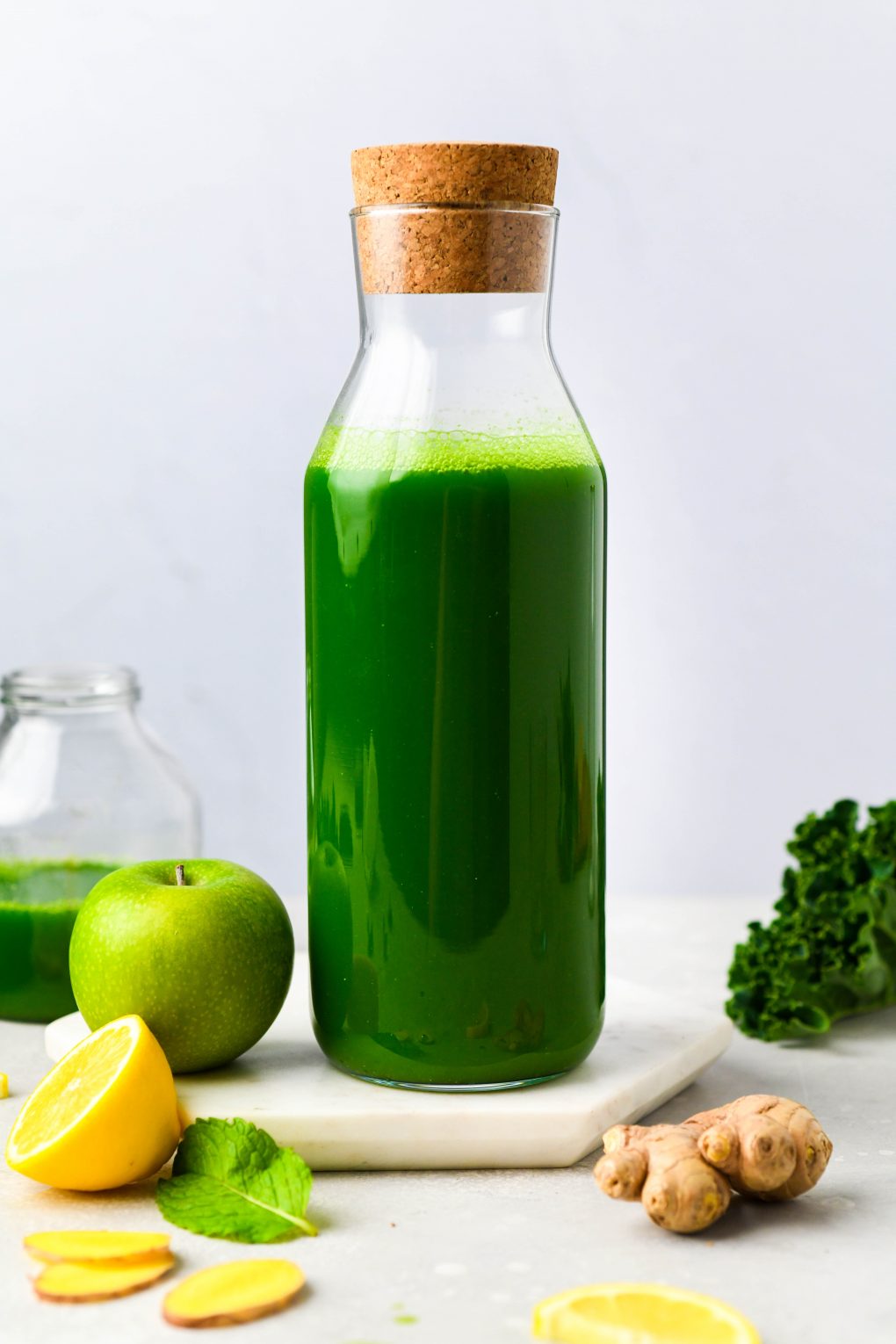 Straight on shot of a carafe filled with green juice. On a light background surrounded by a green apple, a sliced lemon, ginger, fresh mint, and a few kale leaves.