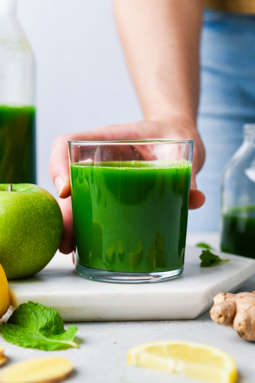 Straight on shot of a hand holding a small glass of green juice. On a light background surrounded by a green apple, a sliced lemon, ginger, fresh mint, and a few kale leaves.