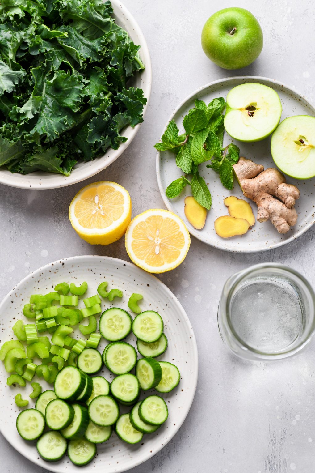 Overhead shot of the various ingredients used to make green juice in a blender. A Large white speckled bowl filled with kale leaves, a small plate with a cut apple, fresh mint, cut ginger, a lemon cut in half, another plate with sliced celery, sliced cucumber, and a mason jar with water. On a light grey background.