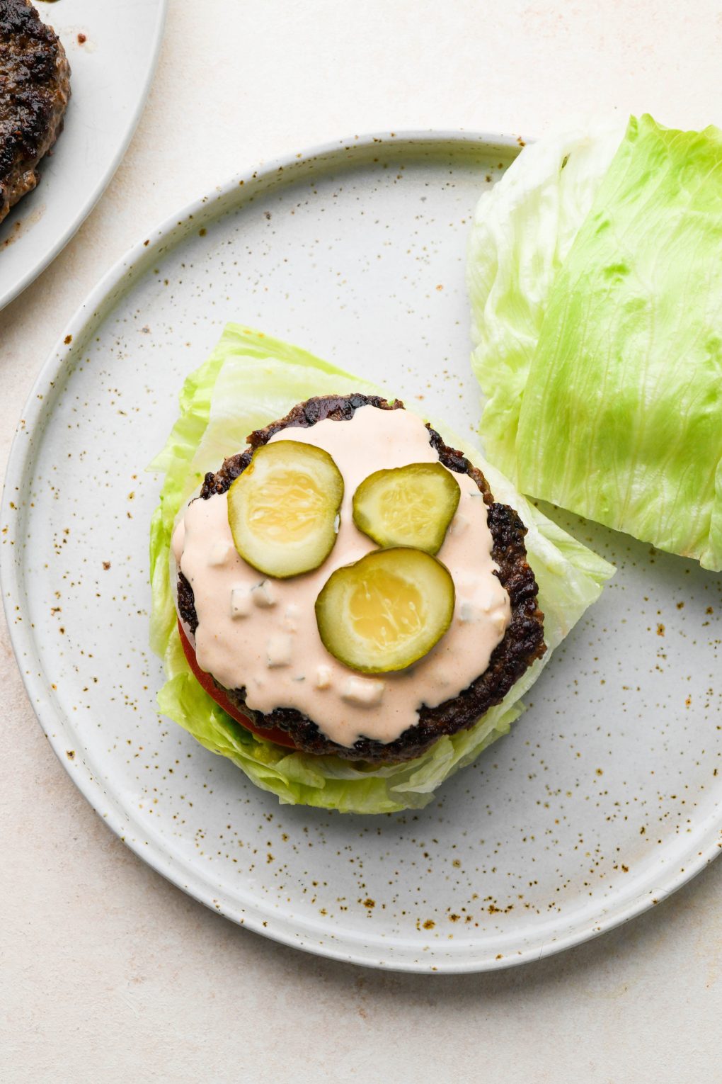 How to make a bunless lettuce wrap burger: lettuce wedges on a plate topped with smashed avocado, tomato, thinly sliced white onion, a burger patty, burger sauce, and sliced pickles.