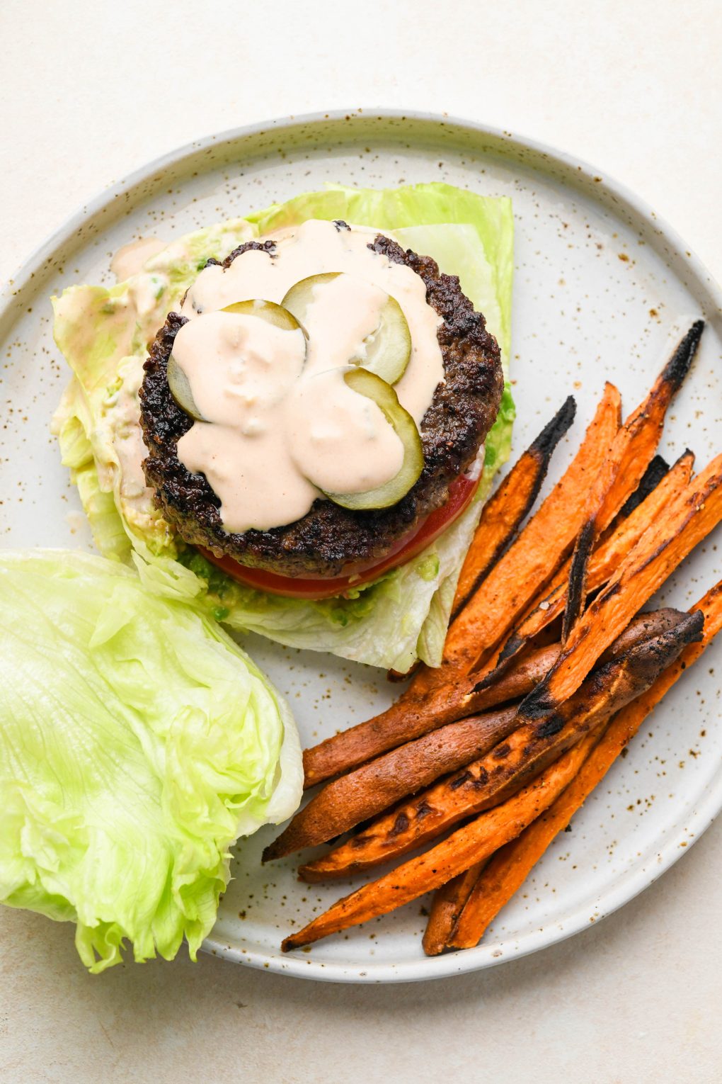 Overhead of a Lettuce Wrap Bunless Burger next to sweet potato fries on a white speckled plate.