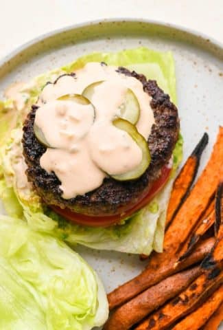 Lettuce wedge on a plate topped with smashed avocado, tomato, thinly sliced white onion, a burger patty, burger sauce, and sliced pickles, next to some sweet potato fries.