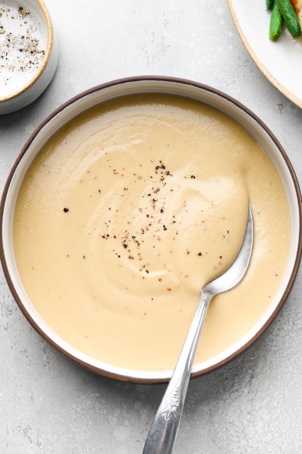 Overhead image of a small bowl of make ahead whole30 gravy seasoned with black pepper with a spoon dipping into it.