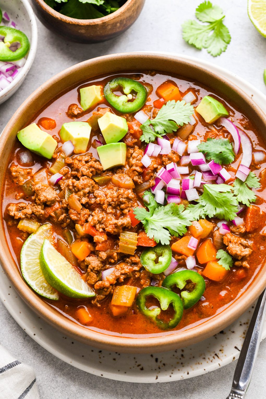 Colorful bowl of beanless Whole30 chili topped with avocado, red onion, cilantro, sliced jalapeno, and lime wedges.