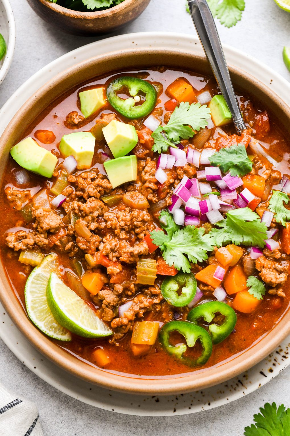 Colorful bowl of beanless Whole30 chili topped with avocado, red onion, cilantro, sliced jalapeno, and lime wedges.