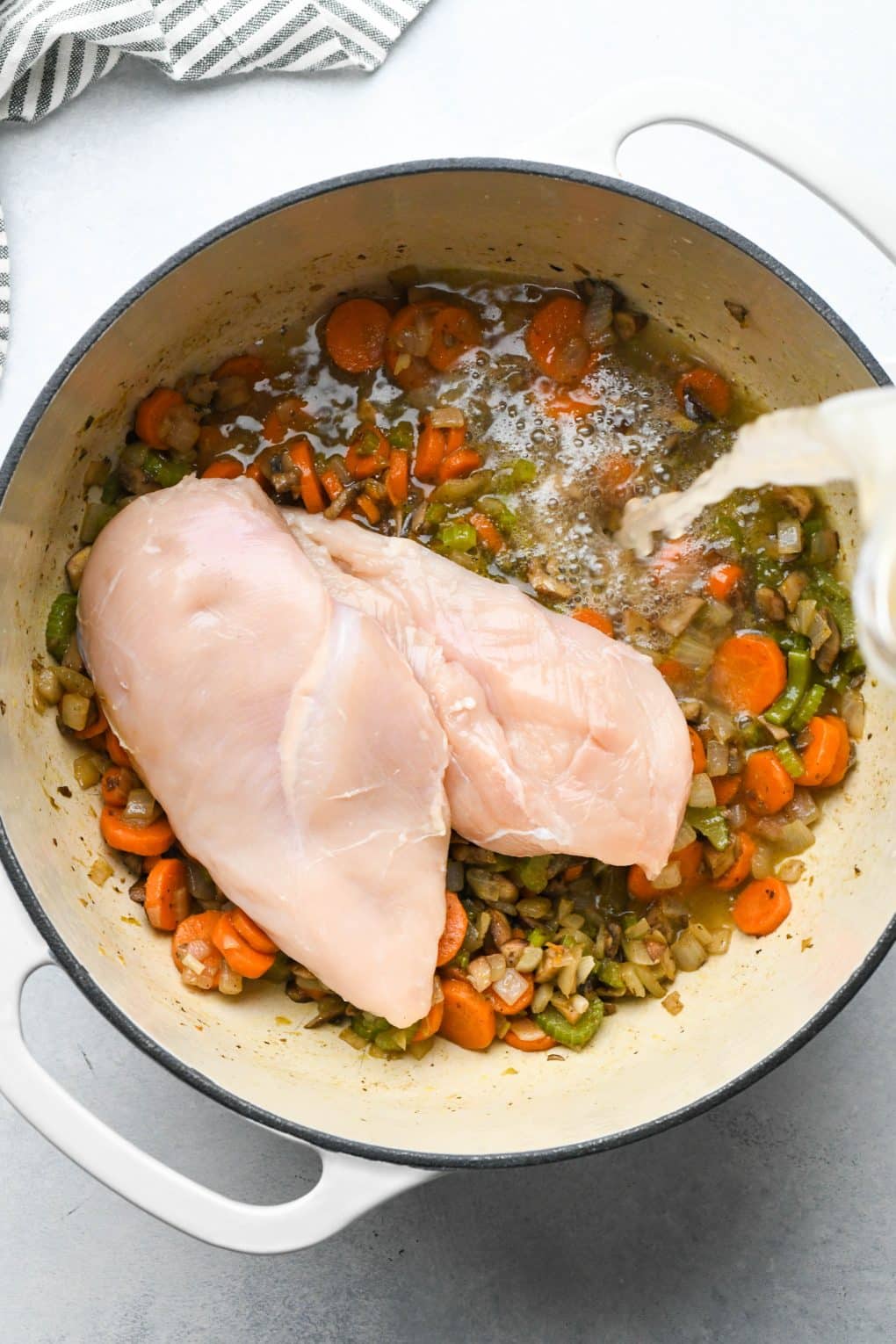How to make Creamy Dairy Free Chicken and Cauliflower Rice Soup: Raw chicken breasts in soup pot with cooked veggies, a hand pouring in the chicken broth.