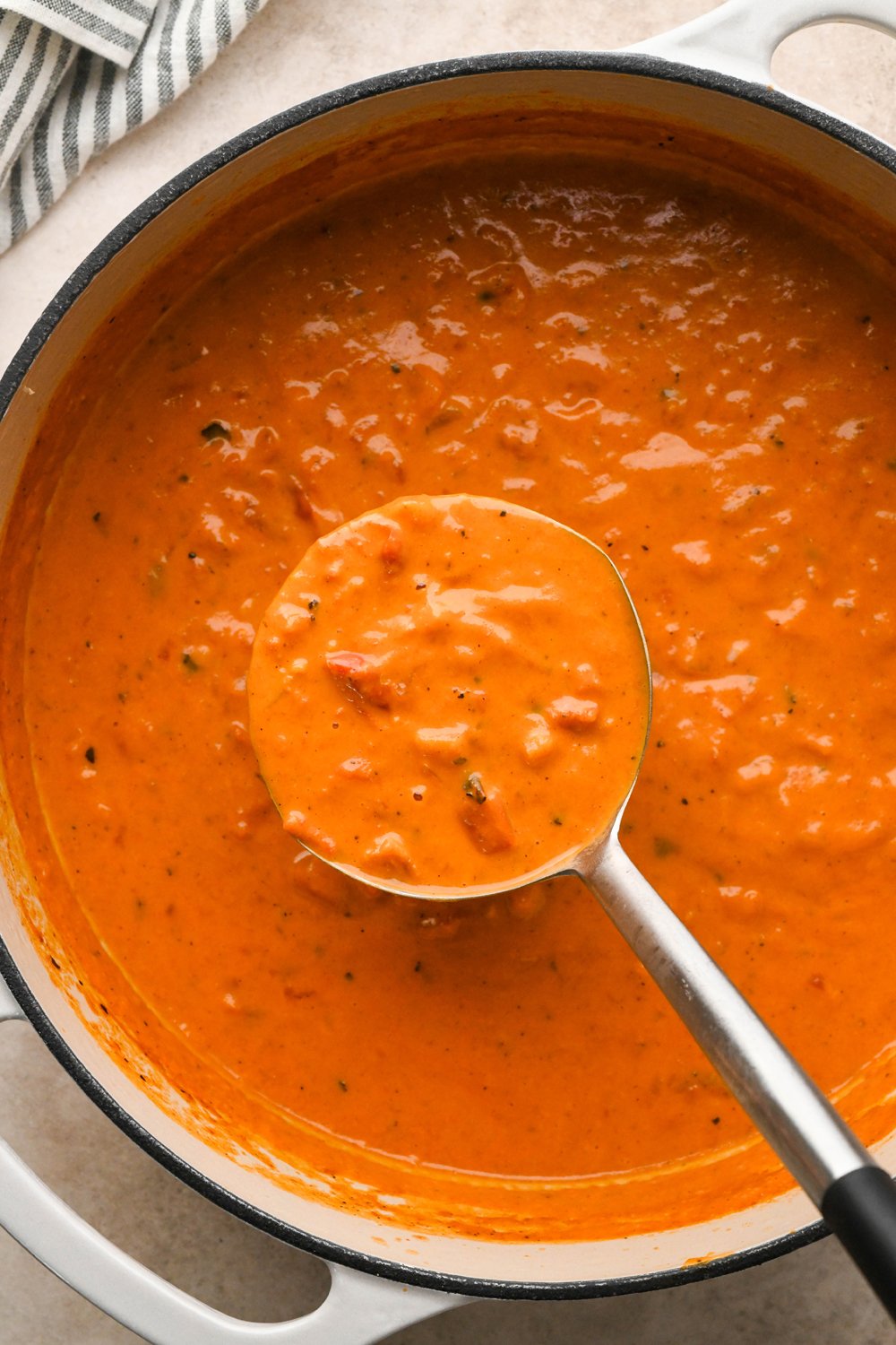 A large creamy white enameled soup pot of dairy free tomato and red pepper soup with a ladle dipping into the soup.