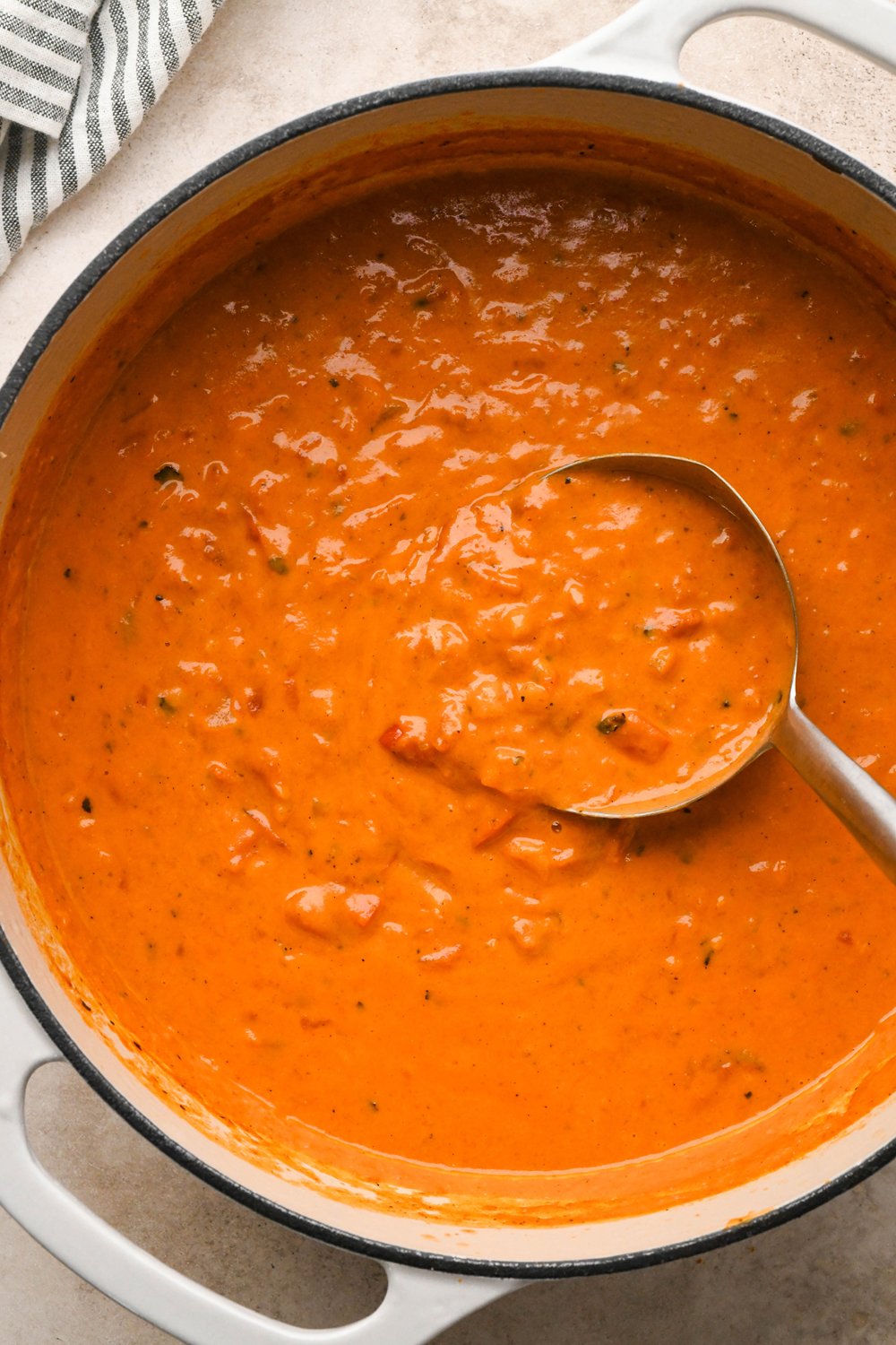 A large creamy white enameled soup pot of dairy free tomato and red pepper soup with a ladle dipping into the soup.