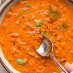 A large ceramic bowl of thick and creamy tomato and red pepper soup, garnished with fennel fronds and fresh basil leaves. A spoon is dipping into the soup to show the luscious texture.